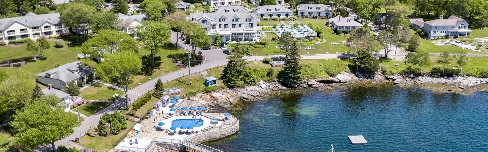 An aerial view of a coastal resort with multiple buildings, a swimming pool, a pier, and a sailboat, surrounded by lush greenery and water.