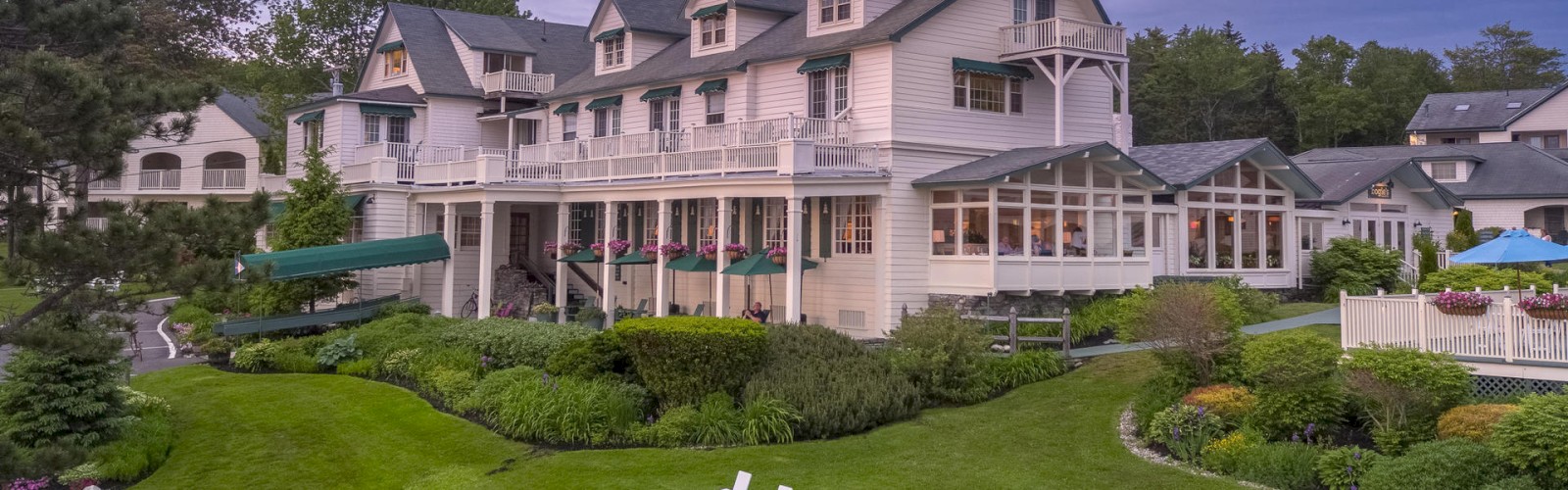 A large, multi-story white house with green roofs sits in a landscaped garden; white chairs are arranged on the lawn, and trees surround the area.