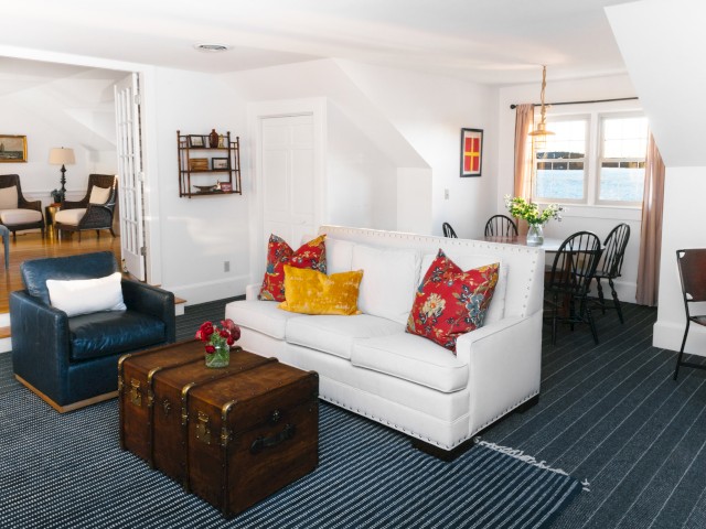 A cozy living room with white sofa, colorful cushions, trunk-style coffee table, and navy armchair, adjacent to a dining area with black chairs.