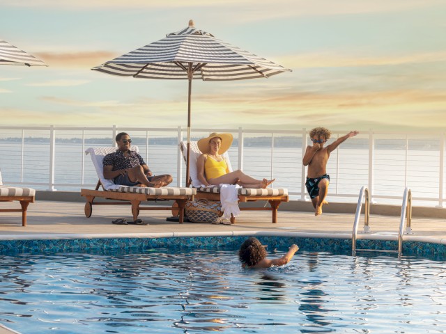 Two adults relax on lounge chairs under umbrellas by a pool, while two children play in the water with an ocean or lake in the background.