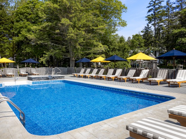 A serene outdoor pool surrounded by lounge chairs with striped cushions and umbrellas in a wooded area, perfect for relaxation and recreation.