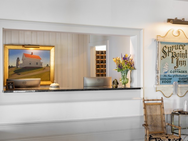This image shows a hotel reception desk with two computers, flowers in a vase, a framed painting, and a vintage sign that says 