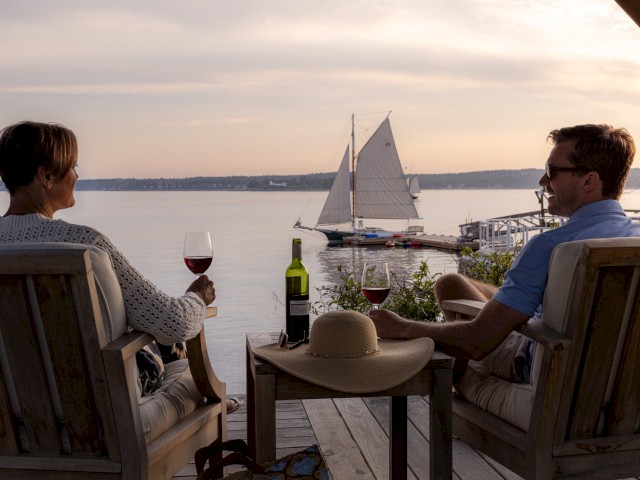 Two people are sitting on a deck by the water, enjoying glasses of wine while a sailboat passes by in the background.