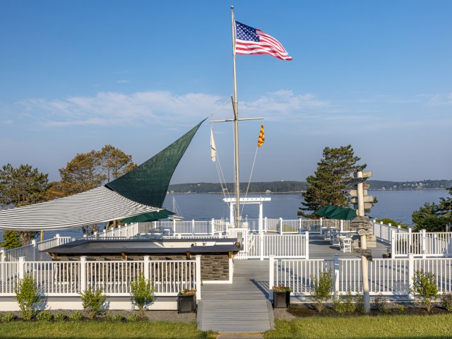 The image shows an outdoor deck area overlooking a lake, with an American flag flying atop a flagpole, surrounded by greenery and boats in the water.
