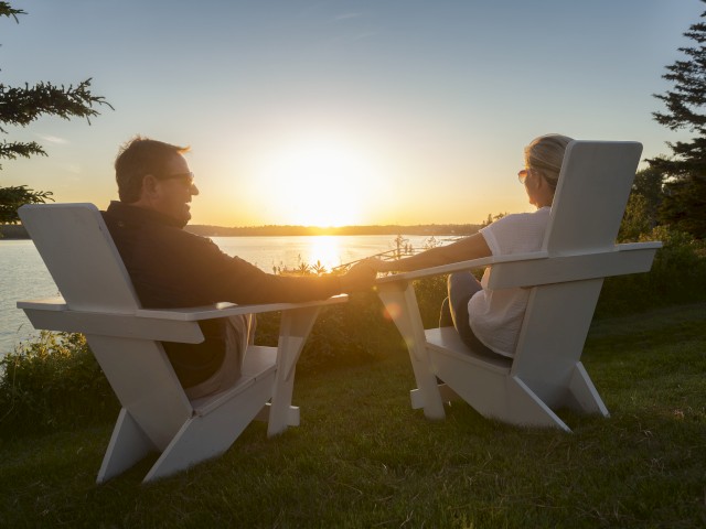 Two people sit on Adirondack chairs by a lake, holding hands and watching the sunset, surrounded by trees and grass, ending the sentence.