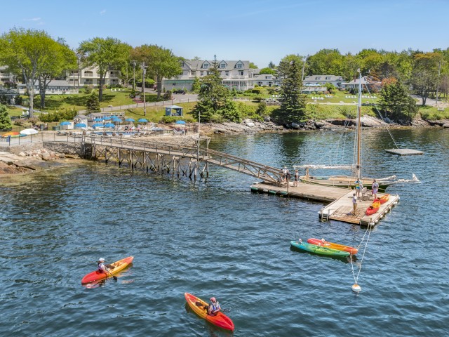 People are kayaking near a dock with a sailboat, houses, and trees in the background, under a clear blue sky.