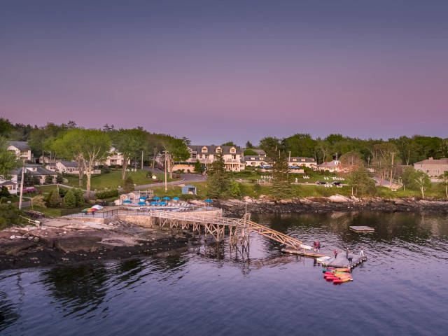 A serene coastal scene at sunset with brightly colored kayaks on the water, houses on the green hillside in the background, and a wooden dock.