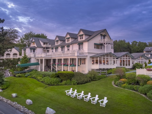 A large, elegant building with multiple gables and a spacious lawn with white chairs arranged in a row, set against a vibrant, cloudy sky.