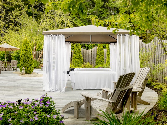 A serene outdoor setting with a canopy-covered massage table, surrounded by greenery, wooden chairs, and flowers, providing a relaxing atmosphere.