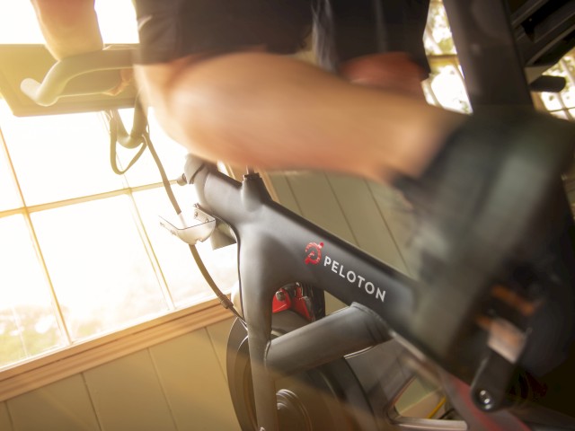 A person is riding a Peloton exercise bike indoors with sunlight filtering through the window in the background, creating a bright atmosphere.