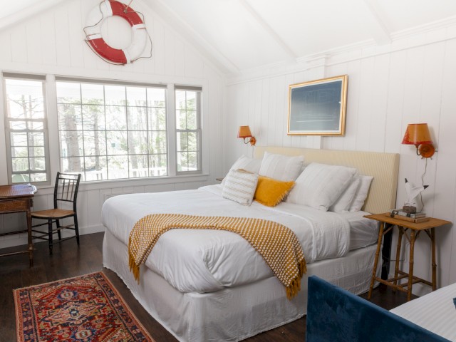 A cozy bedroom with a large bed, side tables, a rug, a desk with a chair, a large window, and a life preserver decoration on the wall.