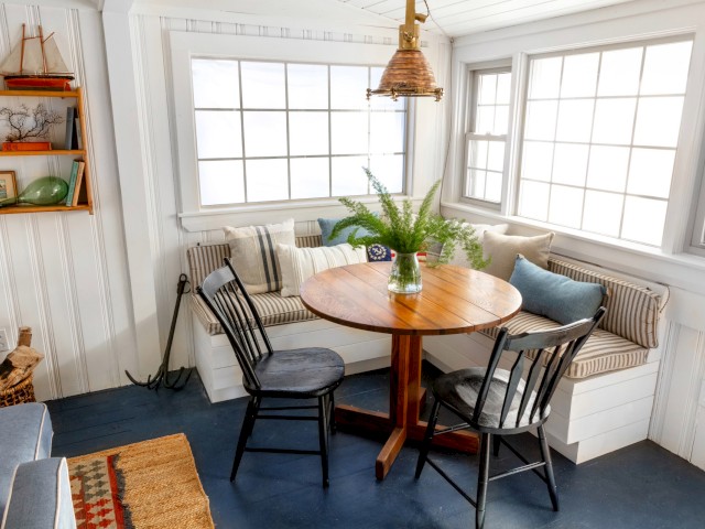 This image depicts a cozy dining nook with a round wooden table, two chairs, cushioned bench seating, and a plant centerpiece near large windows.