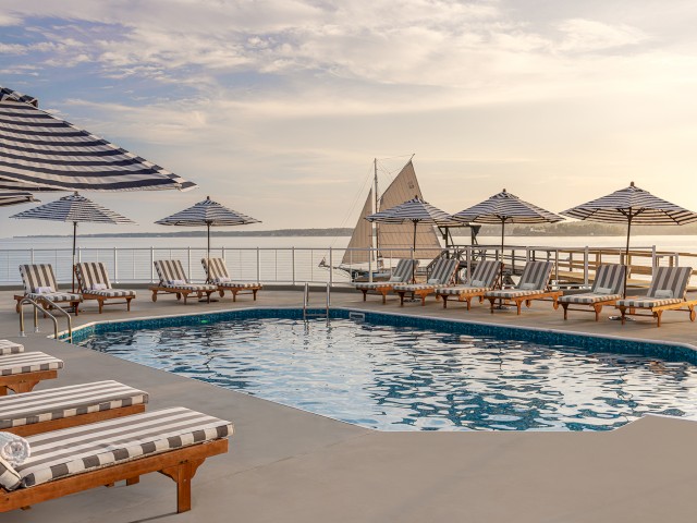 A serene poolside area with striped lounge chairs and umbrellas overlooks a calm body of water with a sailboat in the distance and a picturesque sky.