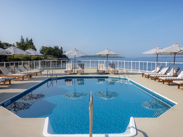 A clear swimming pool is surrounded by lounge chairs with striped cushions and white umbrellas, set against an ocean backdrop under a blue sky.