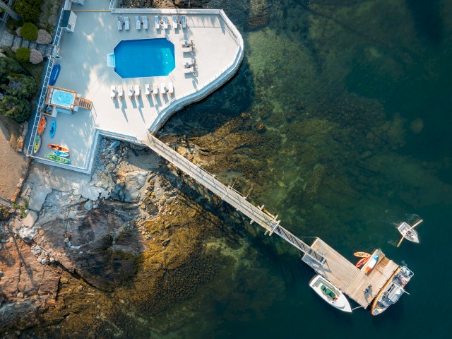 An aerial view of a pool and relaxation area beside a rocky shoreline, connected by a wooden dock to boats moored in clear water.