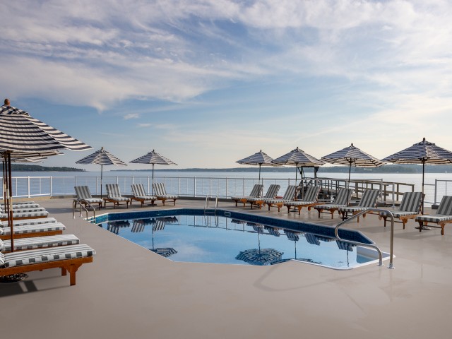 The image shows a serene poolside area with striped loungers and sun umbrellas overlooking a calm body of water under a partly cloudy sky.