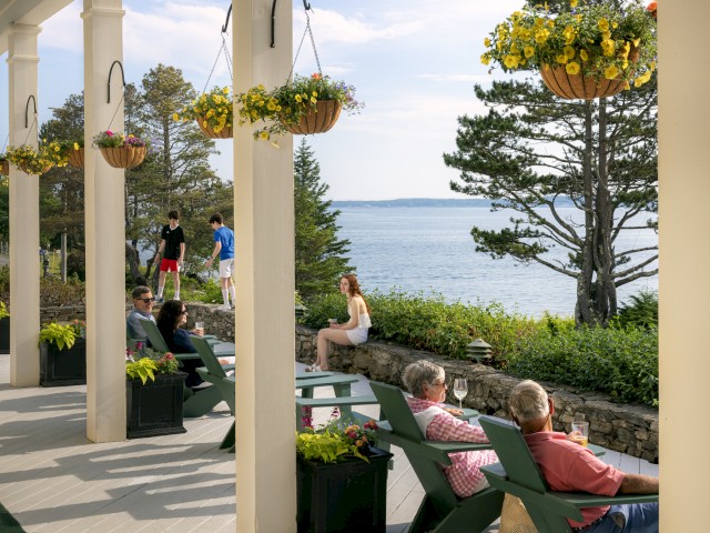 People relax on a porch with flower pots, overlooking a scenic ocean view with trees and a grassy area under a clear sky.