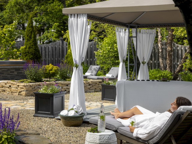 A woman relaxes on a lounger under a canopy in a garden, surrounded by lush greenery, stone accents, and blooming purple flowers.