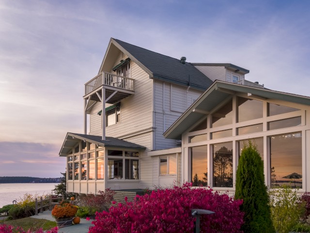 A beautiful lakefront house with large windows, surrounded by colorful bushes and plants, showcasing a peaceful evening sky and water view.