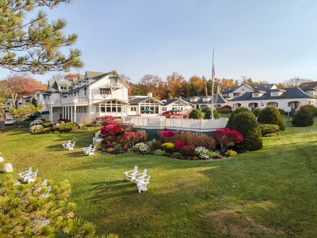 A picturesque garden with vibrant flowers, well-manicured lawns, white chairs, and charming buildings in the background under a clear sky.