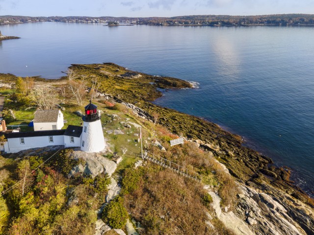 A scenic view of a lighthouse perched on a rocky coastline near calm blue waters, surrounded by greenery and distant coastal town.