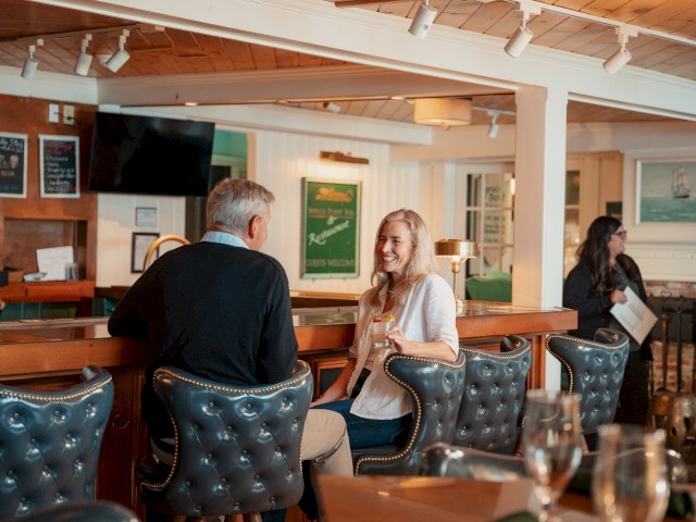 Two people are sitting at a bar, engaged in conversation and smiling, inside a cozy, warmly-lit restaurant or café.
