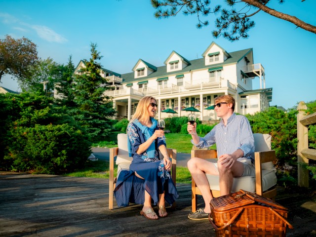 A couple is enjoying drinks outdoors near a large house with green roofs, surrounded by greenery, with a picnic basket beside them.
