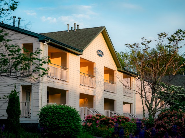 The image shows a two-story white building with balconies, surrounded by trees and bushes, under a blue sky.