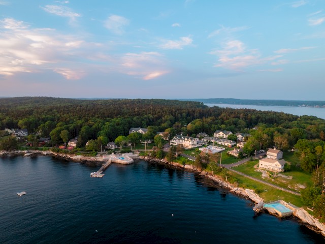Aerial view of a coastal landscape with houses, trees, and a dock by a serene body of water under a blue sky with clouds.