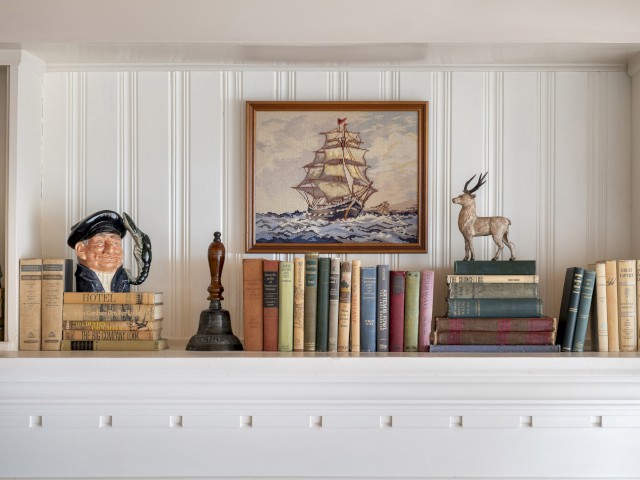 A decorative shelf with books, a ship painting, a bell, a figurine of a deer, and a bust of a sailor.