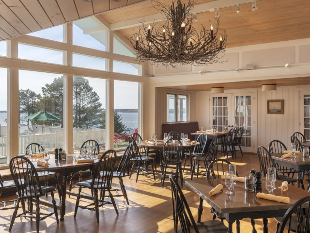 A bright, airy restaurant with wooden tables and chairs, large windows showcasing a scenic seaside view, and a unique chandelier made of branches.