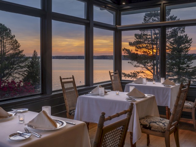 A cozy restaurant with elegant table settings overlooking a beautiful lake at sunset through large windows.