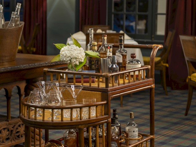 The image shows a wooden bar cart with glassware, bottles, a flower arrangement, and other bar accessories in a vintage room setting.