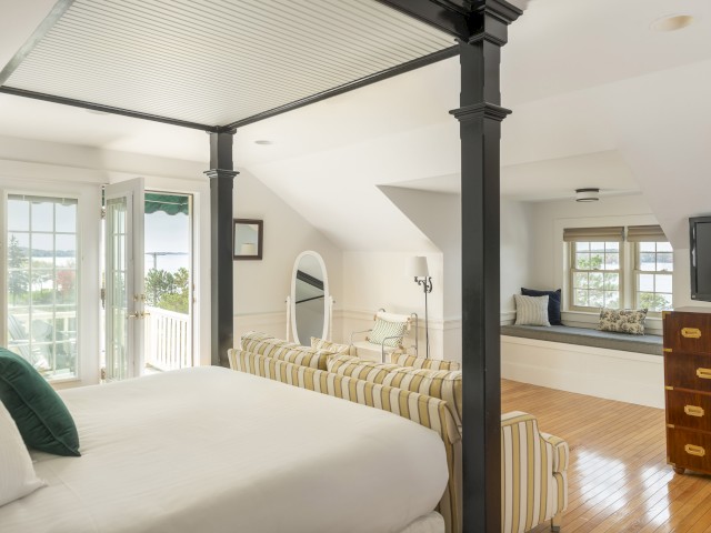 A spacious bedroom with a white bed, striped sofa, large mirror, wooden dresser, TV, and a window seat. French doors open to a balcony.