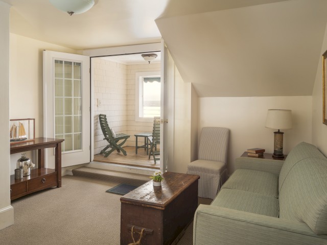 A cozy living room with a sofa, wooden chest, console table, lamp, and doorway leading to a small, enclosed sunroom with two chairs.