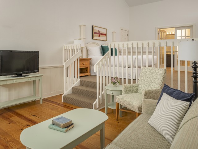 This image shows a cozy living area with a TV, sofa, and armchair, leading up to a raised bedroom area with a bed and nightstand with lamps.