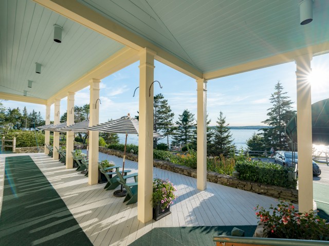 A scenic porch lined with potted plants and chairs overlooks a lush garden and a distant water view under a clear sky with the sun shining.