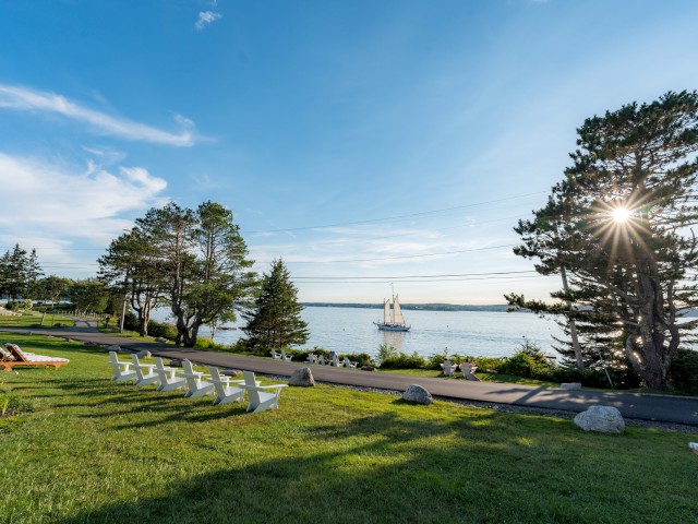 A scenic view of the coastline with Adirondack chairs on a lawn, trees, and a sailing ship in the water under a clear blue sky.