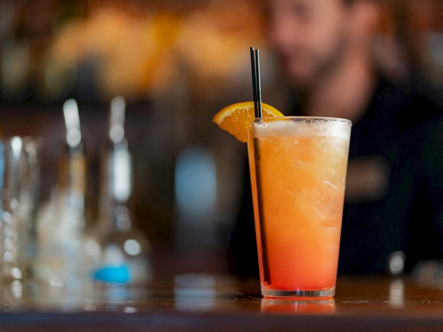 A colorful cocktail with an orange slice garnish and a black straw on a bar counter, with a blurred background of a bartender and bottles.