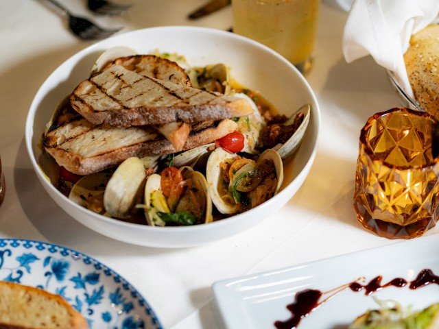 A bowl of seafood pasta topped with grilled bread, surrounded by various other dishes and a glass of amber-colored liquid on a white tablecloth.