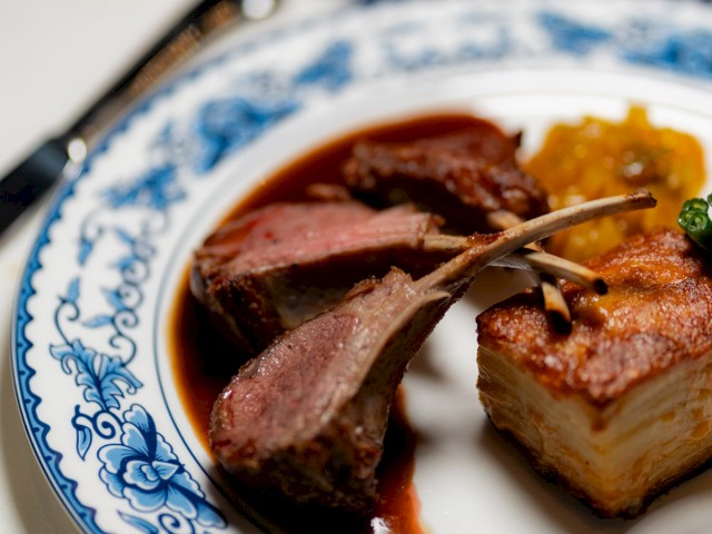 A gourmet dish with lamb chops, a piece of layered meat, and a garnish, served on a decorative plate.
