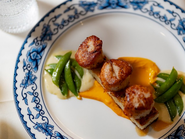 A plate with seared scallops, snap peas, and sauces, garnished beautifully on a white and blue patterned dish, next to a glass of water.