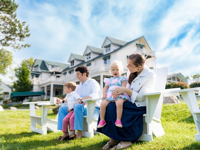 A family sits on white outdoor chairs in front of a large house, with a child on each parent's lap, enjoying a sunny day.