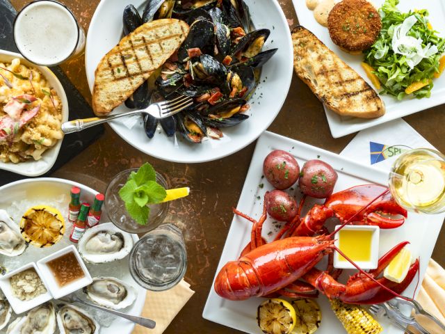 A seafood spread featuring mussels, lobster, grilled bread, oysters, crab cake, potatoes, and drinks, all set on a table.