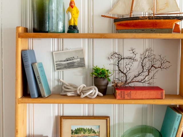 A wooden shelf holds books, bottles, a figure, a model ship, photo, potted plant, branches, framed artwork, rope, and a green glass object.