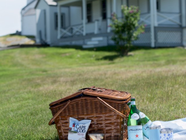 A picnic setup with a basket, snacks, and drinks on a striped blanket in front of a house with a lighthouse in the background, ending the sentence.
