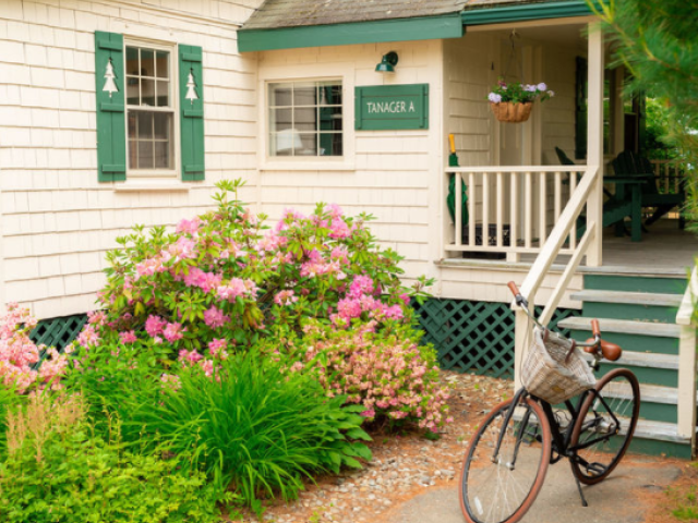A cozy house with green shutters, a small porch, blooming flowers in front, and a bicycle resting against the porch railing ends the sentence.