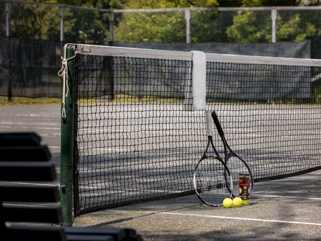 A tennis court with two tennis rackets leaning against the net, along with a canister and three tennis balls.