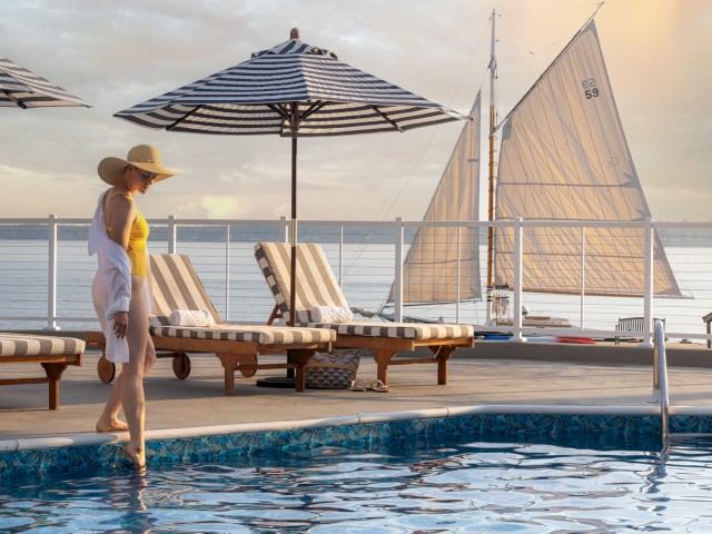 A person in a swimsuit and hat stands by a pool with striped umbrellas, lounge chairs, and a sailboat on the water in the background.