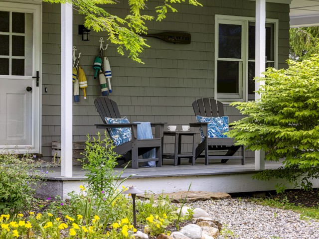 A cozy porch with two chairs, cushions, and a small table is adorned with hanging towels, plants, and greenery in the surrounding area, ending the sentence.